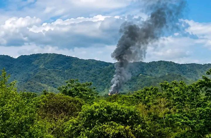 Thick smoke rising from a village area surrounded by hills and forest